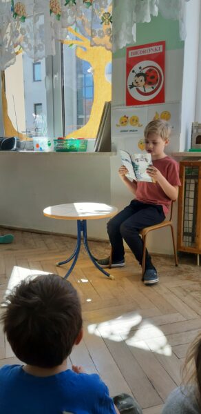 Na krzesełku siedzi chłopiec, w dłoniach trzyma książkę, przed nim siedzą na podłodze dzieci.