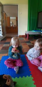 Na matach siedzą dzieci, dziewczynka wącha jabłko.
