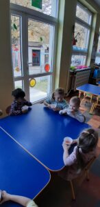 Przy stoliku siedzą dzieci. jedna dziewczynka stoi, wkłada do sokowirówki jabłka.