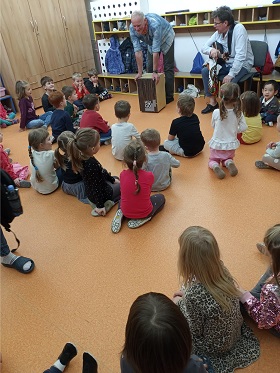 Dzieci siedzą na podłodze i słuchają opowieści prowadzącego; drugi prowadząca trzyma gitarę w ręce
