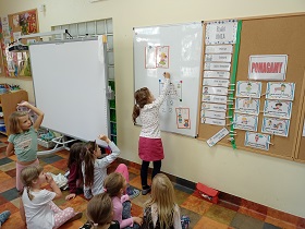Dziewczynka stoi przy tablicy magnetycznej i zapisuje dwójkę markerem, reszta dzieci siedząca na podłodze na nią patrzy.