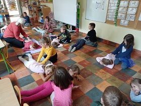 Część dzieci leży na brzuchu na podłodze, część klęczy przy nich i wykonuje im masaż pleców.