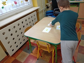 Chłopiec stoi przy stole. Sięga ręką po białą kartkę. Na stole leżą trzy kartki. 