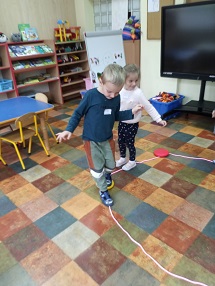 Chłopiec idzie po sznurku ułożonym na podłodze w kształcie koła, próbuje utrzymać równowagę. Za nim idzie dziewczynka.