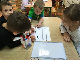 Dzieci siedzą przy stole i malują flamastrami po kartce, na której narysowana jest piramida żywienia.
