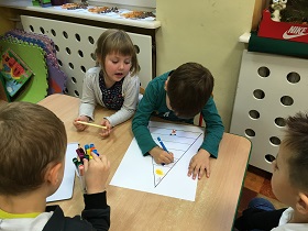 Dzieci siedzą przy stole i malują flamastrami po kartce, na której narysowana jest piramida żywienia.