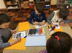 Dzieci pracują przy stołach. Na stole znajdują się kartki, kredki oraz wazony, które dzieci kolorują.