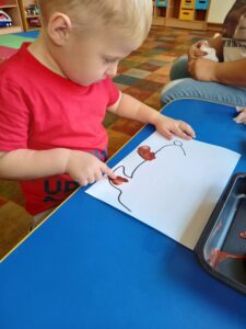 Chłopiec siedzi przy stole, przed nim leży kartka z zarysem kształtu jeża. Dziecko maluje palcem łapy jeża.