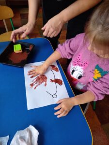 Dziewczynka siedzi przy stole, dłoń umoczoną w brązowej farbie odciska na kartce z narysowanym kształtem jeża.