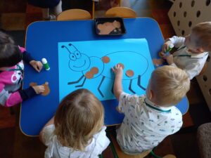 Przy stoliku siedzi czworo dzieci, każde ma klej. Dzieci przyklejają brązowe kółeczka na rysunek mrówki.
