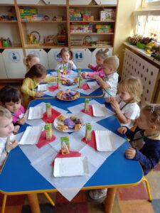 Dziewięcioro dzieci siedzi przy stole. Przed każdym dzieckiem ustawiona jest tacka, sok. Na talerzach ułożone są słodkie przekąski. Dzieci jedzą wybrane przez siebie słodkości.