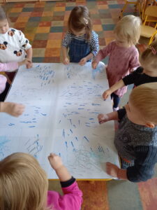 Dzieci stoją wokół stołu na który rozłożony jest duży arkusz papieru. Dzieci uderzają niebieskimi kredkami o papier.