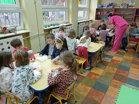 Dzieci siedzą przy stolikach z kartkami przed sobą. Pani w różowym stroju coś im tłumaczy.