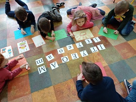 Dzieci leżą i siedzą na podłodze z kartkami papieru i flamastrami. Na środku sali leżą literki na białym tle: I,i,T,t,Y,y,E,e,O,o,A,a,M,m.