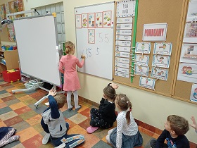 Dziewczynka stoi przy tablicy i piszę na niej mazakiem 5. Na podłodze siedzą dzieci i trzymają rękę w górze.
