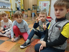 Na podłodze siedzi czwórka dzieci. Jeden chłopiec pokazuje na jednej ręce trzy palce, na drugiej dwa palce.