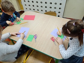 Dzieci odrysowują swoje dłonie na kartkach papieru. Na stoliku leżą kartki, nożyczki i ołówki.