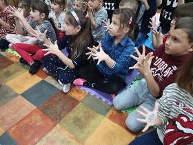 Dzieci siedzą w siadzie skrzyżnym na podłodze. Mają otwarte dłonie i rozsunięte palce.