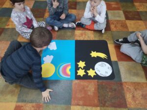 dzieci siedzą na podłodze, przed nimi leży karton podzielony na dwa kolory, czarny i niebieski, dzieci dopasowują elementy do ilustracji