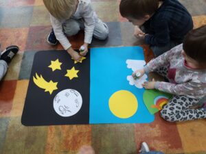 dzieci siedzą na podłodze, przed nimi leży karton podzielony na dwa kolory, czarny i niebieski, dzieci dopasowują elementy do ilustracji