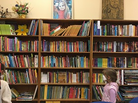 W tle widać regały z książkami. Przed nimi stoją dwie dziewczynki.