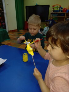 Dwoje dzieci siedzi przy stole, trzyma gwiazdkę na patyku i ją malują żółtą farbą.