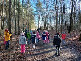 Dzieci stoją w lesie na ścieżce. Przed nimi idzie Pan.