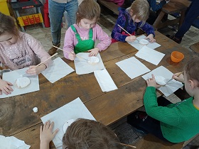 Dzieci siedzą przy drewnianym stole i malują pędzlami na biało drewienka ułożone na ręczniku papierowym.