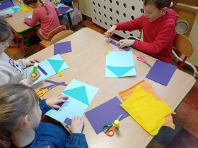 Dzieci siedzą przy stoliku i przyklejają elementy do kartki. Na stole leżą nożyczki, kartki fioletowe i niebieskie oraz kawałki bibuły.
