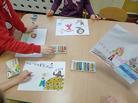 Na stoliku leżą cztery kartki A4 oraz pastele. Dzieci rysują na kartkach różne postaci.