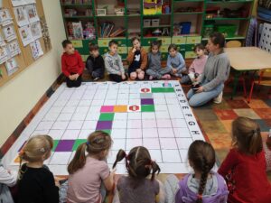 dzieci siedzą na około maty do kodowania, na której ułożona jest gra planszowa z kolorowych kartoników
