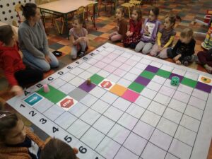 dzieci siedzą na około maty do kodowania, na której ułożona jest gra planszowa z kolorowych kartoników