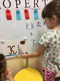 Dziecko stoi przy tablicy magnetycznej. Palcem dotyka literki k. Napisy na tablicy: opert i literki k.