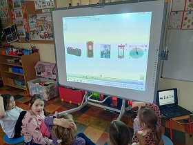 Dzieci siedzą przed tablicą multimedialną i podnoszą jedną rękę do góry. Na tablicy przedstawione są obrazki zegarów. Obok tablicy na krzesełku znajduję się laptop.
