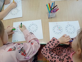 Przy stoliku siedzi trójka dzieci. Dziewczynki mają na swoich kartkach tarczę zegarów, w których rysują elementy flamastrami. 