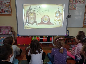 Dzieci siedzą przed tablicą multimedialną. Na tablicy wyświetlany jest film z zegarami.
