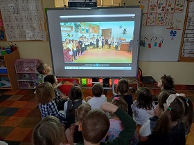 Dzieci siedzą przed tablicą multimedialną i oglądają siebie na ekranie.
