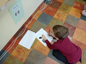 Chłopiec siedzi na podłodze i pisze flamastrem w swojej białej kartce z zadaniem. Patrzy na ścianę, gdzie wisi kartka z napisem kredki.