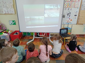 Dzieci siedzą na podłodze przed tablicą multimedialną. Na tablicy widać zdjęcia boiska szkolnego.