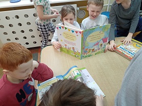 Dzieci siedzą przy stoliku i oglądają książki. Jednak książka stoi na stoliku.