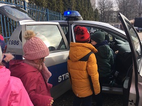 Dzieci stoją przy radiowozie straży miejskiej. Drzwi samochodu i bagażnik są otwarte.