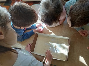 Dziewczynka pędzelkiem przebiera w kaszy mannej, a trójka dzieci się temu przygląda. Jeden chłopiec trzyma karteczkę w dłoniach.