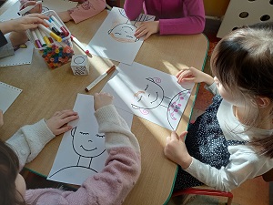 Dziewczynki siedzą przy stole i rysują flamastrami na kartce, na której widniej zarys głowy. Rzucają przy tym papierową kostką.
