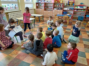 Czwórka dziewczynek przedstawia rysunki w formie książki, dzieciom siedzącym na podłodze. Dzieci są zwrócone głowami do dziewczynek.