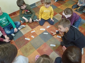 Dzieci siedzą na podłodze w kółku. Przed nimi leżą karteczki z literkami. 