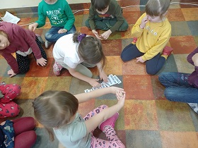 Dzieci siedzą na podłodze w kółku. Dziewczynka układa napisy jeden po drugim.