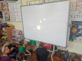 Dzieci siedzą przed tablicą multimedialną i oglądają film. Na tablicy widać planety w układzie słonecznym.