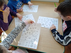 Dzieci siedzą przy stoliku i rozwiązują kartę pracy z planetami, statkami kosmicznymi i gwiazdami.
