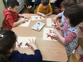 Dzieci siedzą przy stolikach i palcami, brązową farbą wykonują rysunki na kartkach.