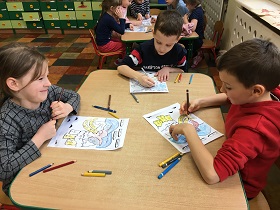 Dzieci siedzą przy stolikach i kolorują kredkami dinozaury.